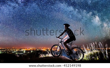 Cyclist riding bike on the top of the mountain. Starry sky, Milky way and night city shining on the background. Picture in the motion. Panorama image