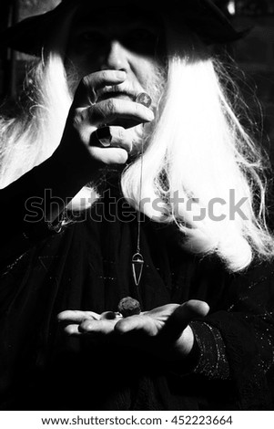 Old man wizard with long gray hair beard in black costume and hat for Halloween holding gem stone and silver pendant for hypnosis on wooden background, black and white