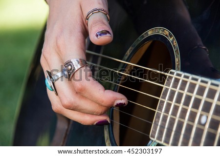 Girl playing guitar in the park in summer. The image is the foreground and fingers and hands are touching the strings of the blue guitar. She is sitting on the grass. 