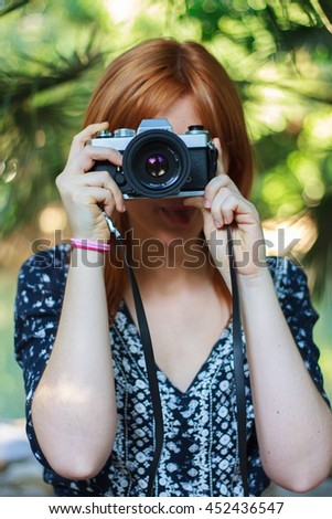 Young girl with orange hair taking pictures with an old camera retro style in a city park with a lake unfocused in the background.  