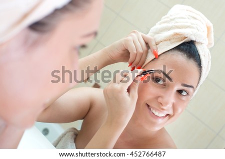 Pretty young woman with towel on head correcting eyebrows with tweezers in front of mirror in bathroom. Beauty and body care concept
