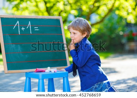Adorable funny little kid boy at blackboard practicing counting and math, outdoor school or nursery. Child having fun with learning. Back to school concept.