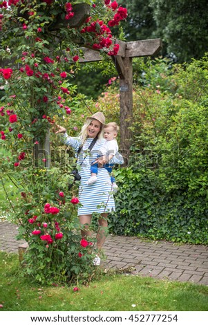 Young mother in a hat holding son on the hands and taking pictures in the park flowers