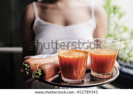 A woman carries a tray of glasses of homemade fresh carrot juice with 2 carrots on the side. Foreground selective focus. Royalty-Free Stock Photo #452789341