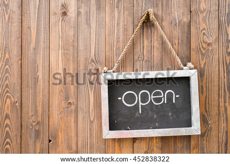 Hand written White 'Open' sign on black board with old steel frame hanging by wicker string on bright brown wooden door