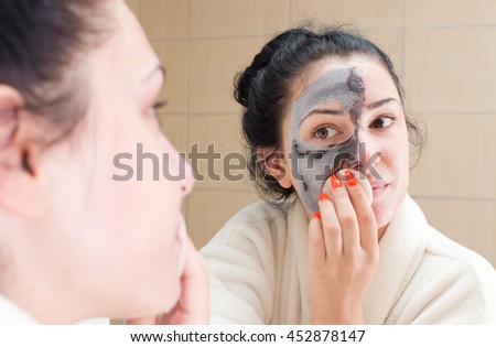 Young pretty woman in bathrobe removing facial mask in front of mirror in bathroom. Skin care and beauty concept