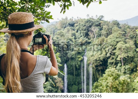 Rear view shot of female tourist photographing a waterfall with her digital camera. Young woman wearing hat taking a picture of water fall in the forest.