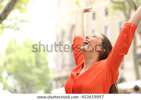 Excited woman wearing orange blouse raising arms outdoors in the street with buildings in the background Royalty-Free Stock Photo #453619897