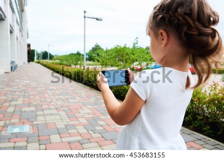 Little preschool girl in white summer dress standing in the street alone hold in hands using smartphone play online game outdoors, new generation addicted with gadgets devices wireless tech concept
