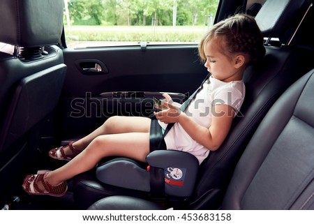 Little girl sitting in a car with a smartphone in hands using applications downloading games watching videos online via internet. New generation gadgets devices modern tech overuse addiction concept
