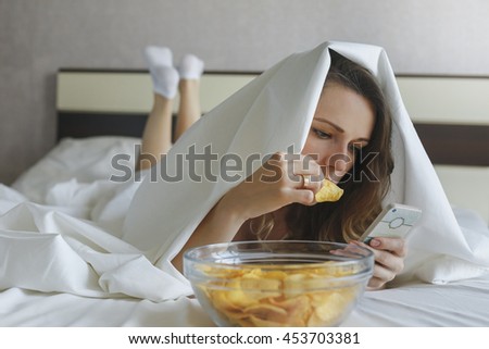 Girl eating chips in bed and watching news in phone Royalty-Free Stock Photo #453703381