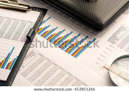 Business accessories (notebook, magnifier, calculator, planchette, tablet, fountain pen, notebook, glasses) and graphics, tables, charts on a wooden office desk. Soft focus