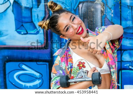 Close up portrait of cute pony tailed teen girl doing thumbs up symbol.Girl holding skate board against colorful graffiti wall.