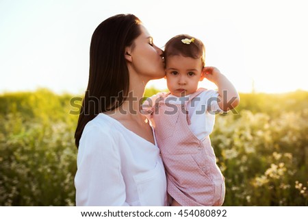 Portrait of tenderness of a pretty brunette mother kiss a daughter, toddler, baby, on sunset, flower field and nature background. Motherhood and childhood.