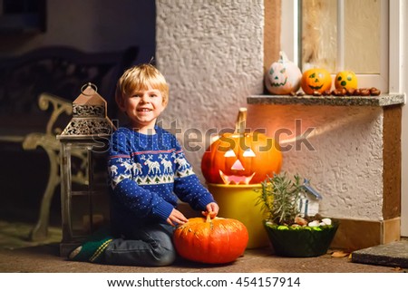 Little kid boy sitting with traditional jack-o-lanterns pumpkins for halloween by the decorated scary door, outdoors. Child having fun and celebrating holiday.