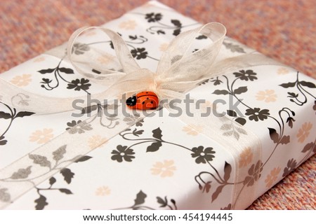 Gift box wrapped with white paper with floral pattern and decorated with ribbon and orange wooden ladybird on pink knitted background. Concept for girls birthday or valentine