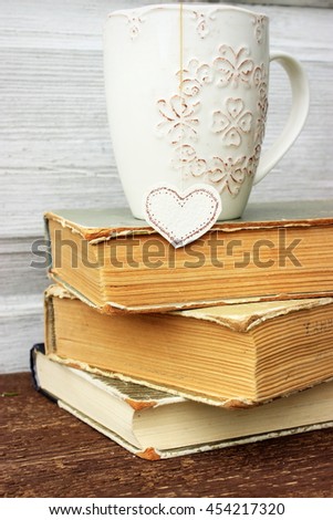 Romantic composition with stack of vintage books with very old paper and covers and cup of hot tasty tea in ceramic cup with label in heart shape. 