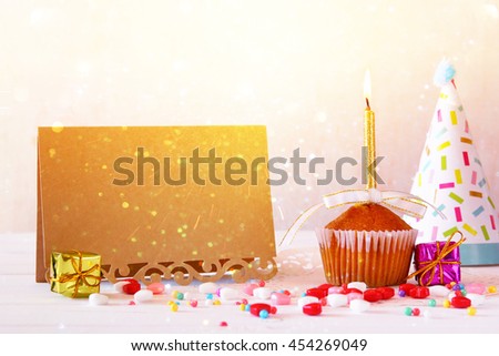 Birthday concept with cupcake and candle next to empty greeting card on wooden table. Glitter overlay