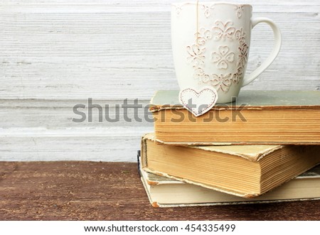 Romantic composition with stack of vintage books with very old paper and covers and cup of hot tasty tea in ceramic cup with label in heart shape on wooden background