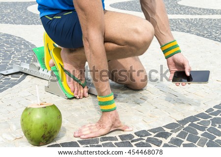 Brazilian athlete wearing flip flops crouching at the start position in running blocks with a coco gelado coconut on the tiles of the Copacabana boardwalk in Rio de Janeiro, Brazil