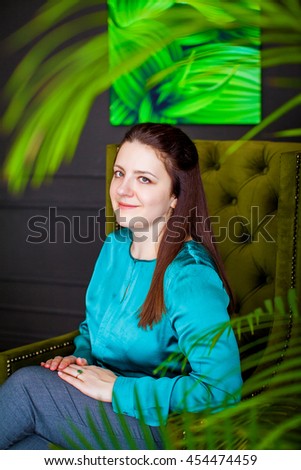 Charming brunette girl sitting in green chair luxurious, on the background of the picture with leaves in a dark room