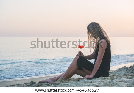 Young blond woman relaxing with glass of rose wine on beach by the sea at sunset. Cleopatra beach, Alanya, Mediterranean region, Turkey.