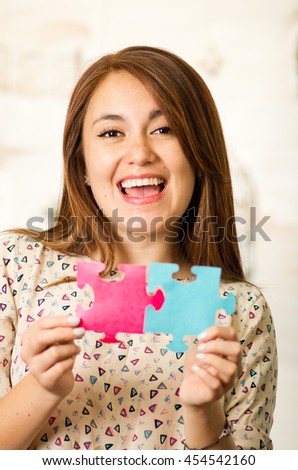 Headshot charming brunette woman holding up big puzzle pieces in pink and blue, smiling happily to camera, white studio background