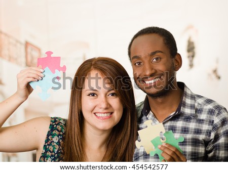 Interracial charming couple embracing friendly, holding up large puzzle pieces and happily interacting having fun, blurry studio background