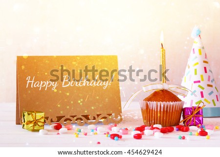 Birthday concept with cupcake and candle next to greeting card on wooden table. Glitter overlay