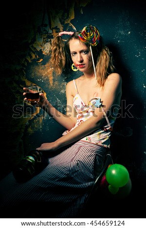Disheveled clown sitting drinking alcohol with a lollipop in her hair.