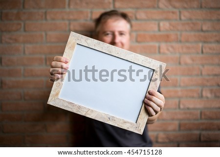 man holds a white wooden frame with blank space, brick wall background - free space for adding custom text or image