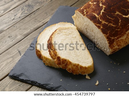 A loaf and slices of crusty white bread on a rustic slate chopping board and wooden table top background