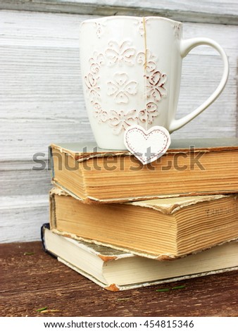 Romantic composition with stack of vintage books with very old paper and covers and cup of hot tasty tea in ceramic cup with label in heart shape on wooden background