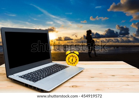 Yellow clock and computer are on the table, blur image of Phuket beach in Thailand at twilight as background.(Dark tone)