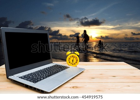 Yellow clock and computer are on the table, blur image of Phuket beach in Thailand at twilight as background.(Dark tone)
