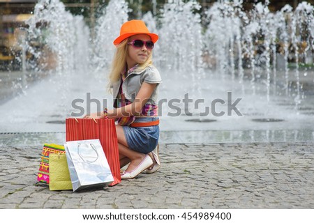 Elegant Charming cute little girl in fashionable clothes and sunglasses, orange hat, sitting with full shopping bags. Looking to side. Shopping bags on pavement. Fountain in background.