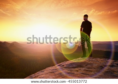 Thinking hiker in black on rocky peak. Wonderful daybreak. Ginger hair man in black at the end of sharp cliff above deep valley. Strong lens defect, reflection