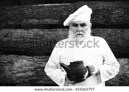 bearded man cook chef in uniform and hat with long beard on serious face holding iron old tea kettle sunny day outdoor on wooden background, black and white