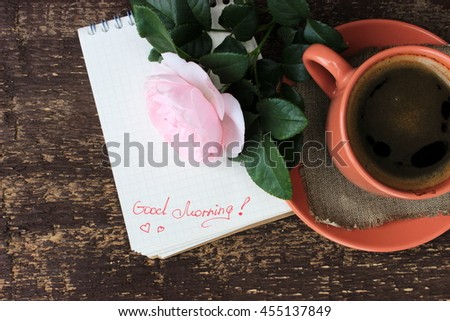 Old brown wooden background with cup of black coffee decorated with linen napkin, notebook with "good morning" text and beautiful pink rose. Romantic rustic design. Copy space. Concept for love theme