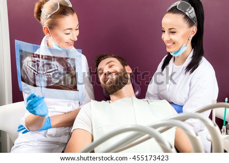 Female doctor and her smiling assistant showing teeth x-ray to male patient in dental hospital. Handsome men with beard sitting in medical chair and looking at the picture.