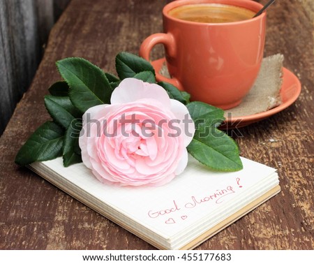 Old brown wooden background with cup of black coffee decorated with linen napkin, notebook with "good morning" text and beautiful pink rose. Romantic rustic design. Copy space. Concept for love theme