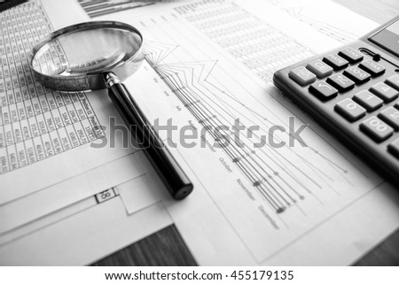 Business accessories (notebook, calculator, planchette, tablet, fountain pen, glasses) and graphics, tables, charts on white sheets on office desk. Soft focus. Black and white.