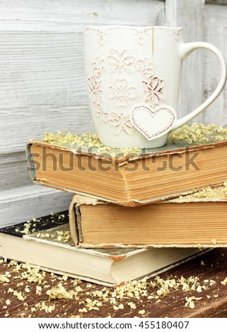 Romantic composition with stack of vintage books with very old paper and covers and cup of hot tasty tea in ceramic cup with label in heart shape on wooden background with petals of dry hydrangea