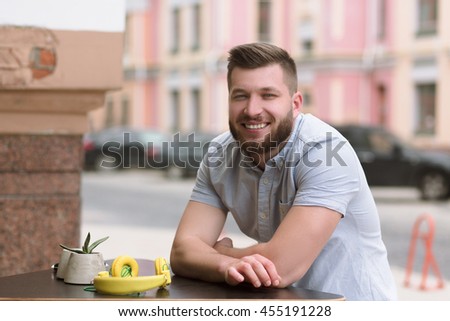 Picture of happy smiling handsome man sitting in outdoor vegan cafe and looking at camera. Bearded man waiting for his order or friends.