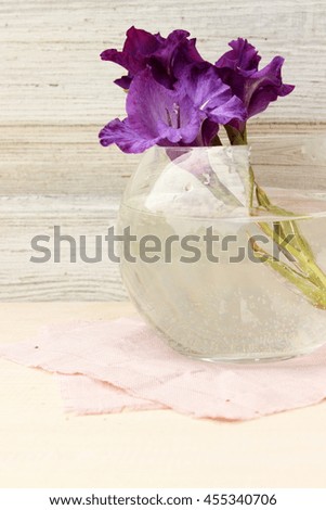 Single flower of purple gladiolus in round glass vase on old white wooden background. Floral decor elements. Concept for romantic greeting card for birthday, valentine, mothers day or weeding