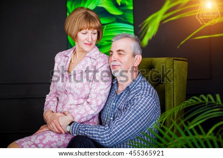 
Old happy retired husband and wife. Grandfather sits in luxurious green chair, grandmother in pink dress sitting on his lap. On the background of tropical plants, in stylish dark room, with picture.