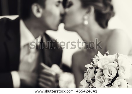 Wedding bouquet lies on the frontground of a black and white picture of kissing newlyweds