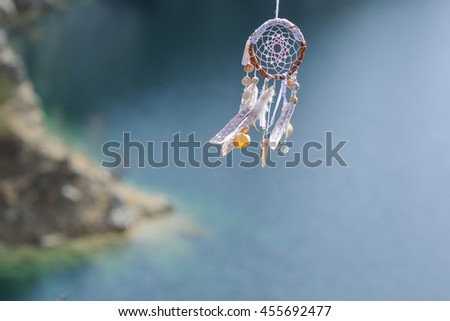 Handmade pink native american dream catcher on background of rocks and lake. Tribal elements, feathers, shells, lace
