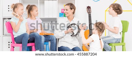 Young woman playing the tambourine among four kids sitting on varicoloured plastic chairs