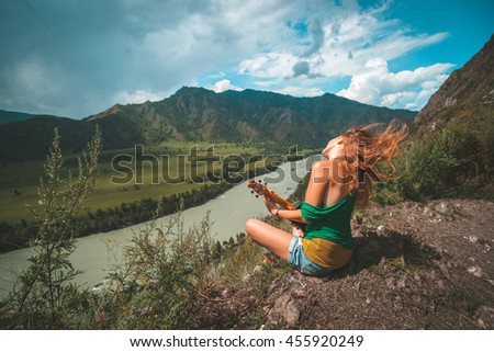 Young red hair woman play ukulele on the hill's summit. Mountain river landscape. 